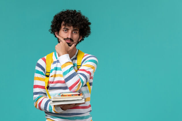 抱身穿条纹衬衫 背着黄色背包 拿着书 在蓝色墙上思考的男生的正面图男人思考男