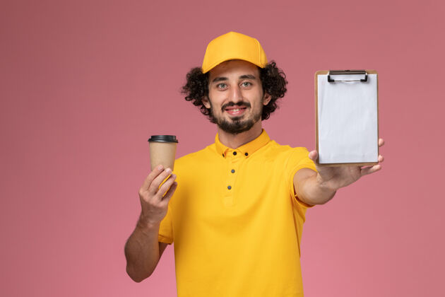 记事本正面图：身穿黄色制服 披风的男性快递员手持咖啡杯和粉色墙上的记事本男士工作手持