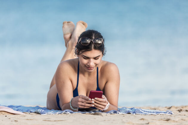 休息在沙滩上用智能手机的女人泳装放松消遣