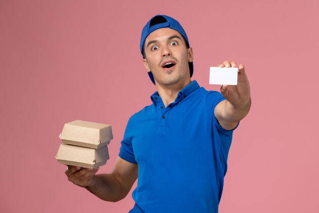 制服正面图：身穿蓝色制服披肩的年轻男性快递员拿着小包裹 浅粉色墙上有卡片服务专业微笑