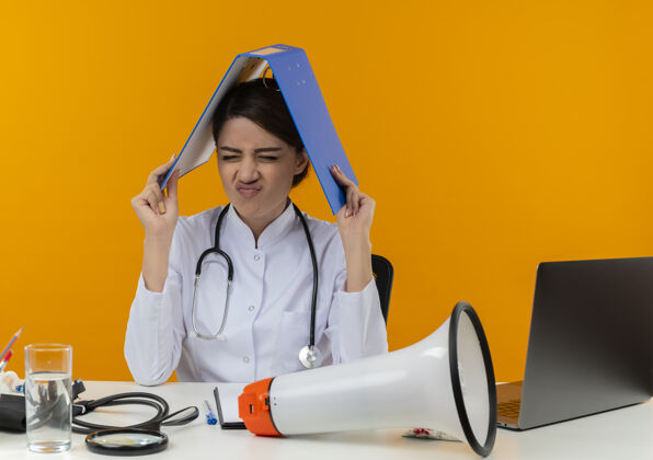 文件夹年轻的女医生闭着眼睛 穿着医用长袍 戴着听诊器 坐在办公桌上 用电脑和医疗工具 头上盖着文件夹 背景是黄色的封面医疗长袍